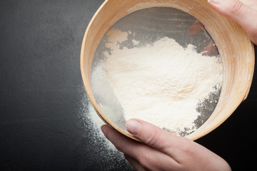 Female hands sift flour on a black background, top view.
