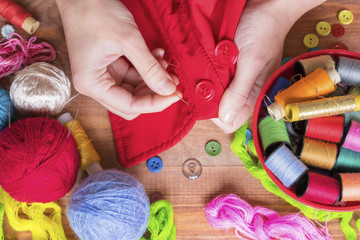 Woman`s hand holding a needle and colorful sewing and knitting supplies on the wooden table, bowl with bobbins, knitting yarn and buttons.