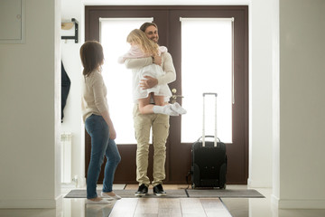Happy father arrived home returning after business trip with baggage, daddy missed little daughter holding in arms hugging girl while wife standing in hall, family reunion, welcome back dad concept