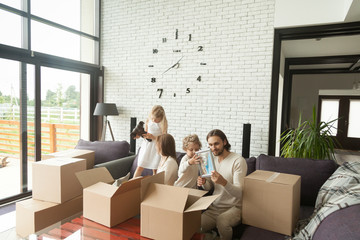 Young happy family with kids unpacking boxes together sitting on sofa in modern living room of country house moving settling in new home, children helping parents to pack stuff move out relocating