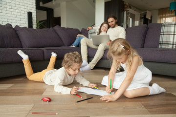 Children little boy and girl having fun on warm floor, boy playing toy car, girl drawing with colored pencils, parents using laptop at home, family leisure together in living room, underfloor heating