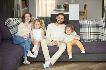 Happy smiling family of four sitting on sofa looking at camera, young couple with kids boy and girl relaxing on couch at home, parents with children posing in living room, mortgage loan, insurance