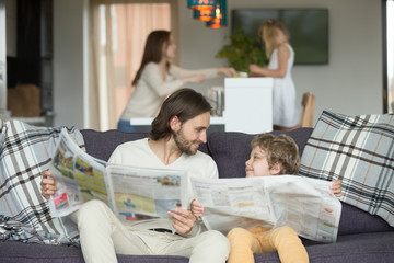 Happy dad and son reading newspapers together on couch, father with little boy looking at each other holding paper news, funny kid copying imitating daddy sitting at home on sofa, intelligent child