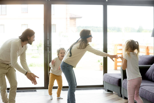 Blindfolded mother playing hide and seek game with father and kids hiding clapping hands at home, happy family having fun together on leisure in living room, parents children activities on weekend