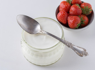 Fototapeta na wymiar White yogurt in glass bowl with spoon on top and starwberries behind on white background.