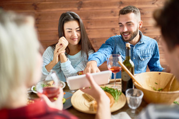 Portrait of young Asian woman and modern man looking at smartphone screen discussing photos while having dinner with friends sitting at big table during holiday celebration.