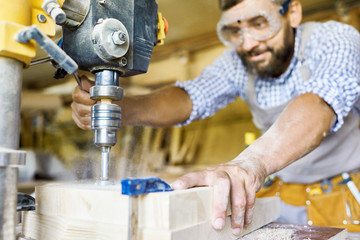 Portrait of mature bearded carpenter wearing protective goggles using drilling machine for piece of wood  in modern workshop, focus on foreground