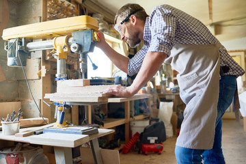 Portrait of  bearded carpenter wearing protective goggles using drilling machine for piece of wood  in modern workshop