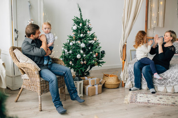 Obraz premium Young happy family of four near christmas tree. Mother with daughter on bed and father with son on armchair. Happy new year concept.