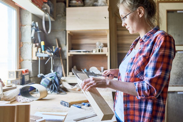 Side view portrait of modern female carpenter measuring wooden part making furniture over working table in workshop