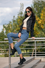 fashionable young woman in rock style clothes, black leather jacket, blue jeans, tights in a grid under battered jeans