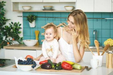 Young mother smiling, cooking and playing with her baby daughter in a modern kitchen. Using phone. Healthy food concept. Working at home.