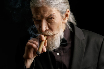 Stylish old man smoking cigar. Aged male in black suit in studio.