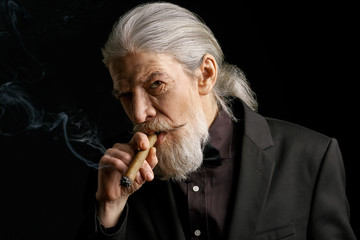 Stylish aged man with long grey hair smoking cigar. Portrait of wrinkled male with cigar on black...