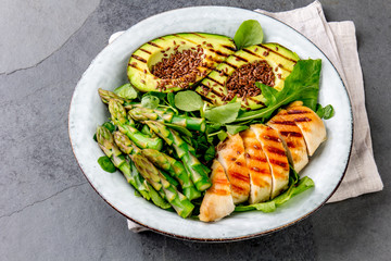Healthy grilled chicken, grilled avocado and asparagus salad with linen seeds. Balanced lunch in bowl. Gray slate background. Top view