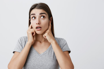 I don't hear anything. Body language. Close up of young attractive caucasian girl with long hair in casual gray t-shirt plugging ears with fingers, looking aside with raised eyebrows and surprised