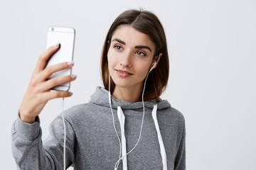 Modern lifestyle. Close up of beautiful young brunette caucasian girl in casual hoodie talking with boyfriend with video on smartphone, wearing headphones, looking in phone screen with happy face