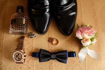 Wedding groom accessories on chair: black shoes, wrist watch, perfume bottle, bow tie near wedding rings