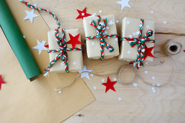 The Christmas gifts packed into a beige kraft paper, strewed with red, white paper stars on the wooden light plane.