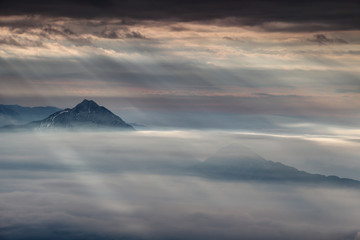 Curtain of sun rays shining through dark gray clouds illuminates two conical mountains in autumn morning mist, Storzic and Tolsti vrh peaks, Kamnik Savinja Alps, Gorenjska, Carniola, Slovenia, Europe 