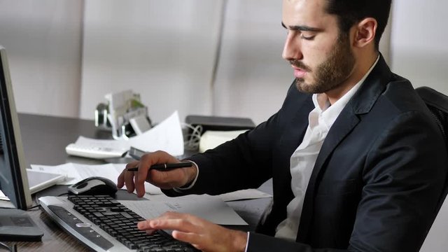 Tired bored young businessman sitting at his desk in front of his computer with his chin resting on his hand, sleepy