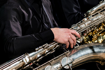 A musician in black clothes sits and holds a baritone saxophone with his right hand. For...