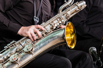The baritone saxophone lies on the knee of the musician in a black shirt and trousers. The right hand lies on a wooden wind instrument.