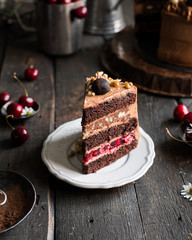 A piece of chocolate cake with cherries and nuts is decorated with mini-cakes, chocolate cheese cream and nuts on a wooden dark background. Fresh cherry and chamomile flowers