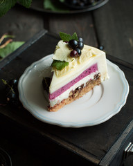 A modern mousse cake with a crusty nut layer, a layer of black wild currant and a mousse of white chocolate and yogurt. The cake is decorated with fresh currant berries and leaves of white chocolate