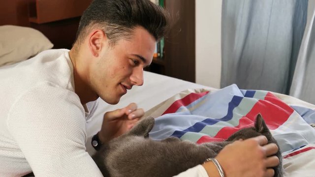 Handsome Young Animal-Lover Man on a Bed, Hugging and Cuddling his Gray Domestic Cat Pet.
