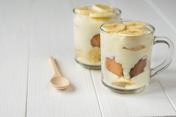 Two glass cups with banana pudding and wooden spoon on a white table. Milk and banana dessert.