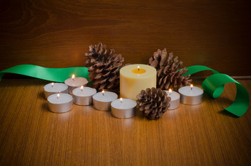 Yellow & white candles with pine cones & green Christmas ribbon over teak wood table and wall background at night time with copy space for text decoration, warm xmas or special event celebration