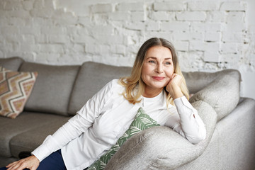 Indoor portrait of attractive happy 60 year old senior woman with pleasant smile relaxing on grey...