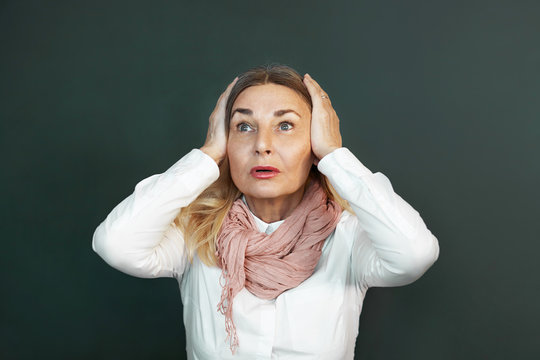 Isolated shot of emotional mature 60 year old Caucasian female wearing white blouse and pink scarf having frustrated look, covering ears with both hands, can't stand unpleasant sound or noise