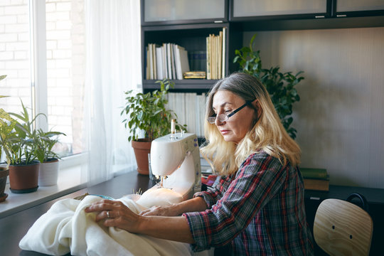 Clothing, fashion design and needlework concept. Blonde mature female dressmaker on retirement mending white dress, working from home, using sewing machine, having serious concentrated expression