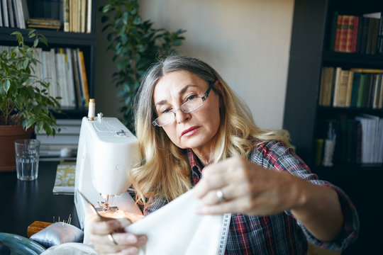 Picture of focused serious middle aged European woman sewing, using stitching machine at home, sitting at her workplace. Elderly female dressmaker holding fabric, scissors and measuring tape