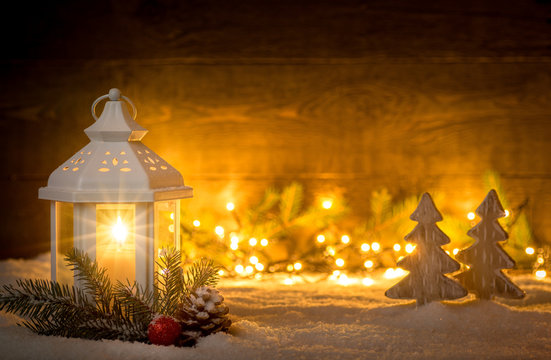 Christmas scene with a lantern, trees, fir branch and blurred lights in front of an illuminated dark wooden board as copy space