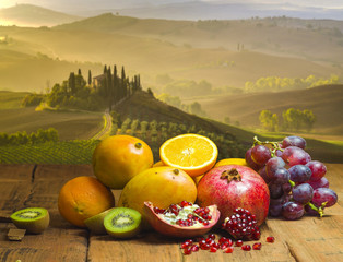 Obrazy na Szkle  owoce na drewnianym stole, w tle pejzaż włoskiej Toskanii