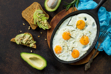 Scrambled eggs on a cast iron skillet and avocado sandwich with fresh sliced avocado and pine nuts on a dark slate or stone background. Top view.