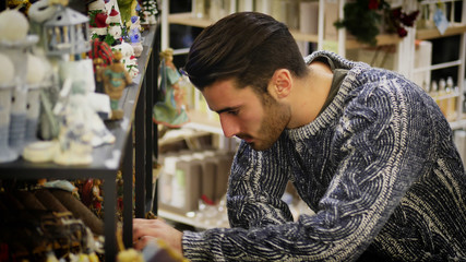 Handsome young man in jumper standing near shelf in shop and looking at goods. 