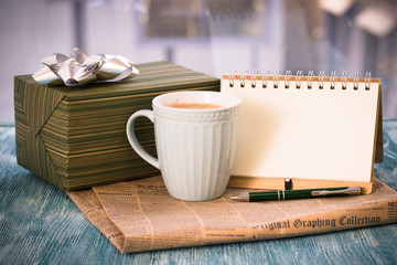 Festive still life with a cappuccino cup, gift box, notebook with pen, newspaper on a turquoise wooden table, country background on a sunny day