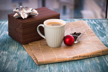 Festive still life with a cappuccino cup, gift brown box, vase with coffee beans, red Christmas-tree ball, newspaper on a turquoise wooden table, country background on a sunny day