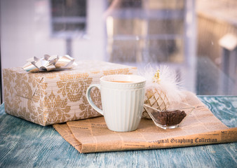 Festive still life with cappuccino cup, gift golden box with bow, vase with coffee beans, shiny Christmas tree ball, newspaper on turquoise wooden table, country background on a sunny day