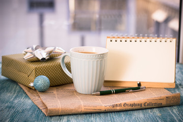 Festive still-life with cappuccino cup, gift box with bow, blue Christmas-tree ball, notebook with pen, newspaper on turquoise wooden table, country background on a sunny day