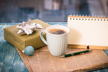 Fototapeta na wymiar Festive still-life with cappuccino cup, gift box with bow, blue Christmas-tree ball, notebook with pen, newspaper on turquoise wooden table, country background on a sunny day