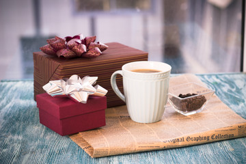 Festive still life with a cappuccino cup, two gift boxes with bows, a vase with coffee beans, a newspaper on a turquoise wooden table, a country background on a sunny day