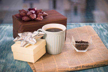 Festive still life with a cup of freshly brewed coffee, two gifts in dark and golden boxes, a vase with coffee beans, a newspaper on a turquoise wooden table, a country background on a sunny day