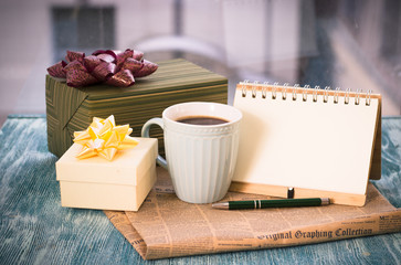 Festive still life with a cup of freshly brewed coffee, two gifts in green and white boxes, a notebook with a pen, a newspaper on a turquoise wooden table, a country background on a sunny day