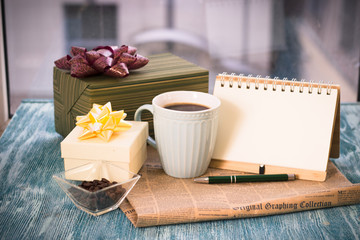 Festive still life with a cup of freshly brewed coffee, two gifts in green and white boxes, a vase with coffee grains, a notebook with a pen, a newspaper on a turquoise wooden table, a country