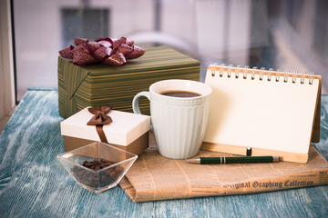 Festive still life with a cup of freshly brewed coffee, two gifts in green and light boxes, a vase with coffee beans, a notebook with a pen, a newspaper on a turquoise wooden table, a country